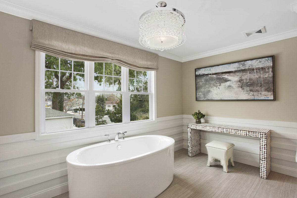 Spacious luxury bathroom with free standing tub, white wainscoting, and bright chandelier